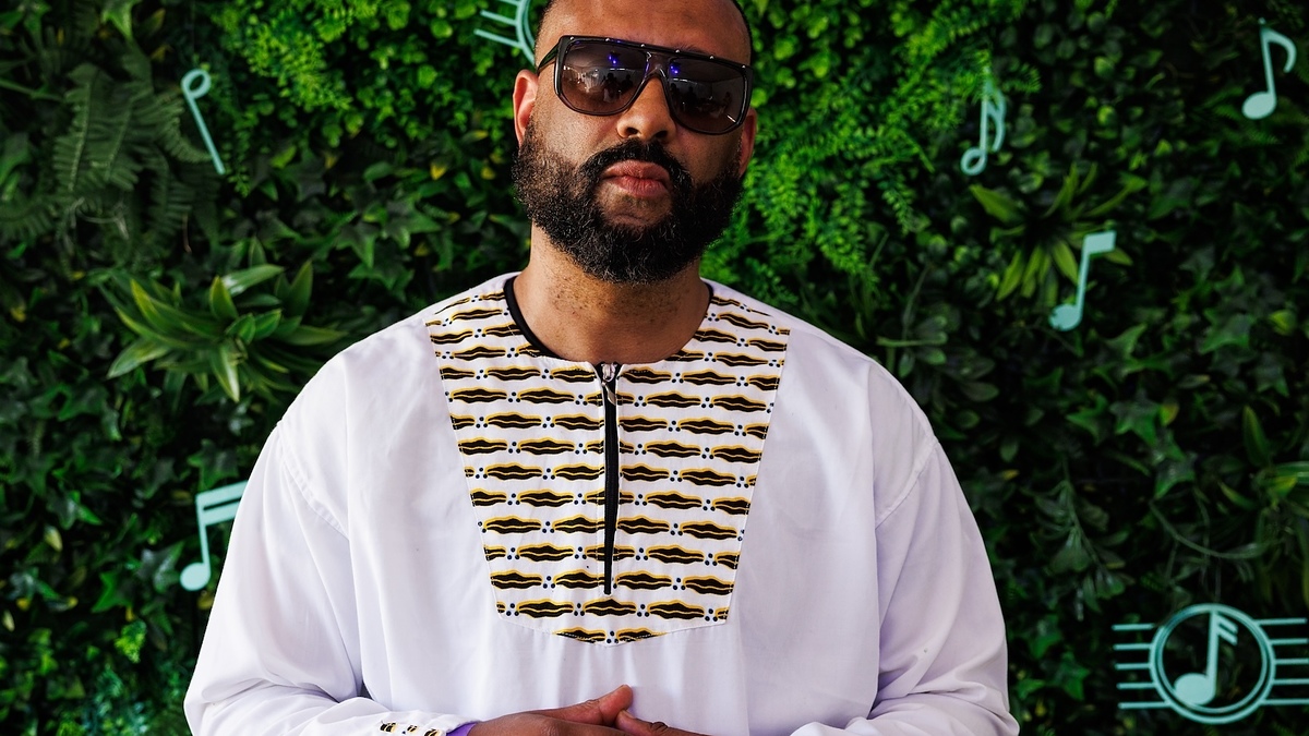 Man in embroidered shirt stands with crossed hands against a leafy backdrop