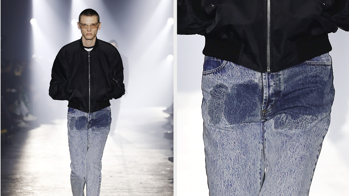 Model on runway in black jacket and faded jeans with wet stain in groin. Close-up shows focus on urine-like stain on denim.