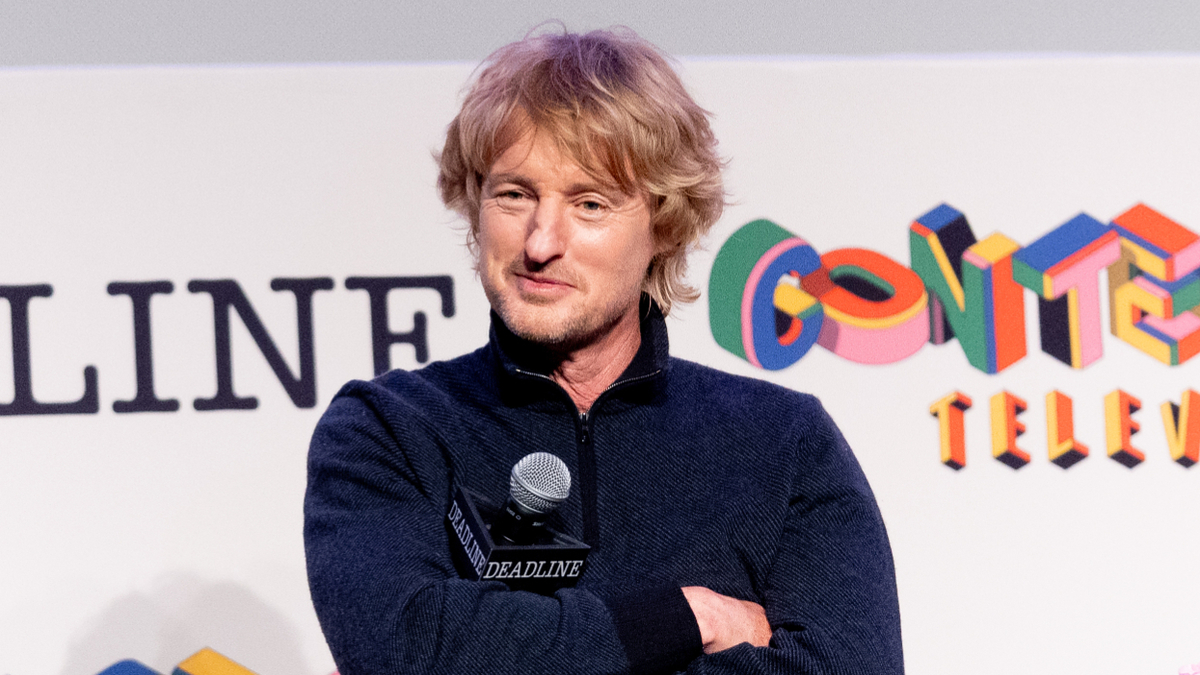 Owen Wilson at an event with microphone, casual attire, speaking onstage with logos in the background