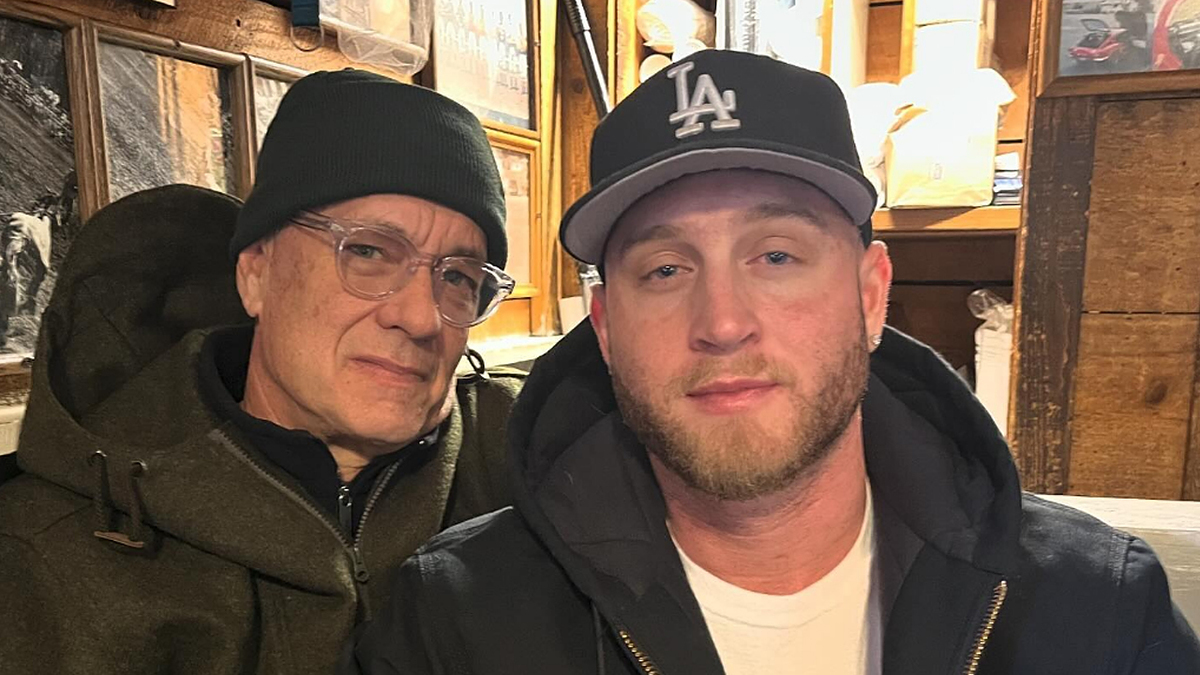 Tom Hanks and Chet Hanks wearing casual jackets and hats, posing together indoors with a rustic, wood-paneled background