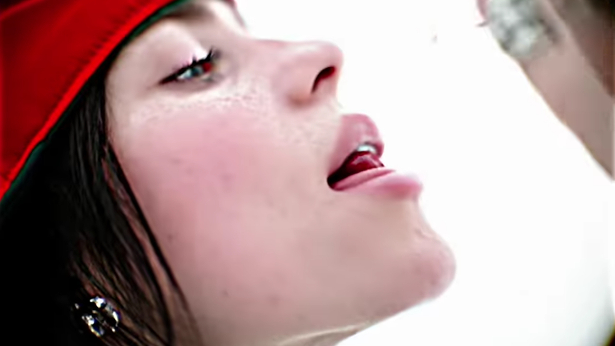 Profile close-up of a woman, likely a musician, wearing a red cap and earrings, sticking her tongue out towards another person whose face is not visible