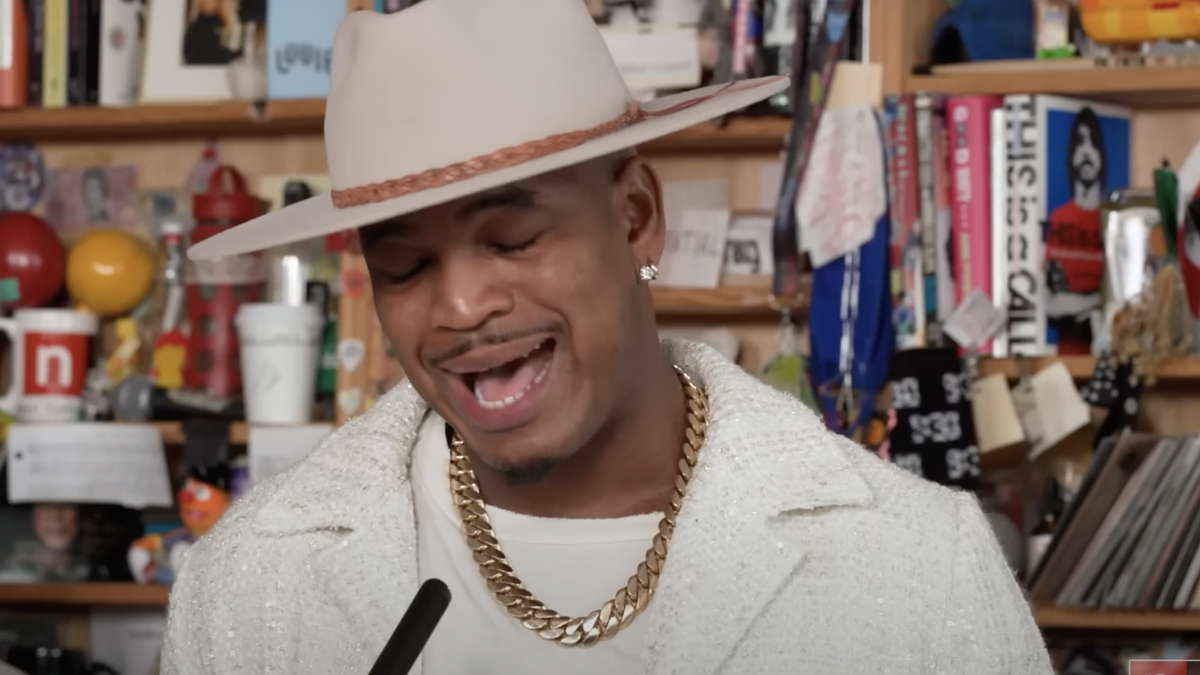 Man in wide-brimmed hat and chain necklace performing at a microphone with shelves in the background