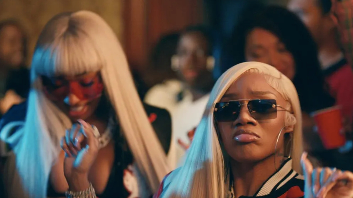 Two women wearing sunglasses and fashionable outfits with a blurred group in the background, in a music-related setting