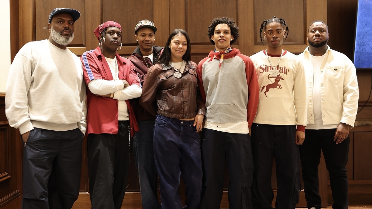 Group of musicians posing together, some wearing casual sweaters and hoodies, with two in front wearing neck scarves