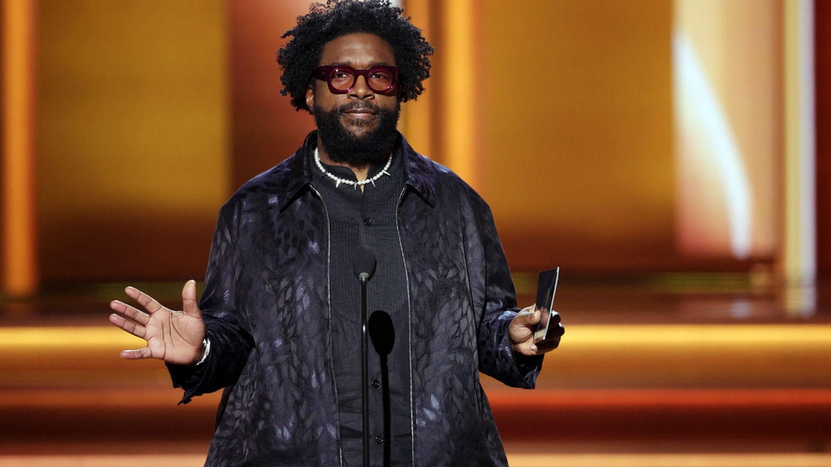 Man in patterned jacket with glasses and necklace standing with microphone on stage