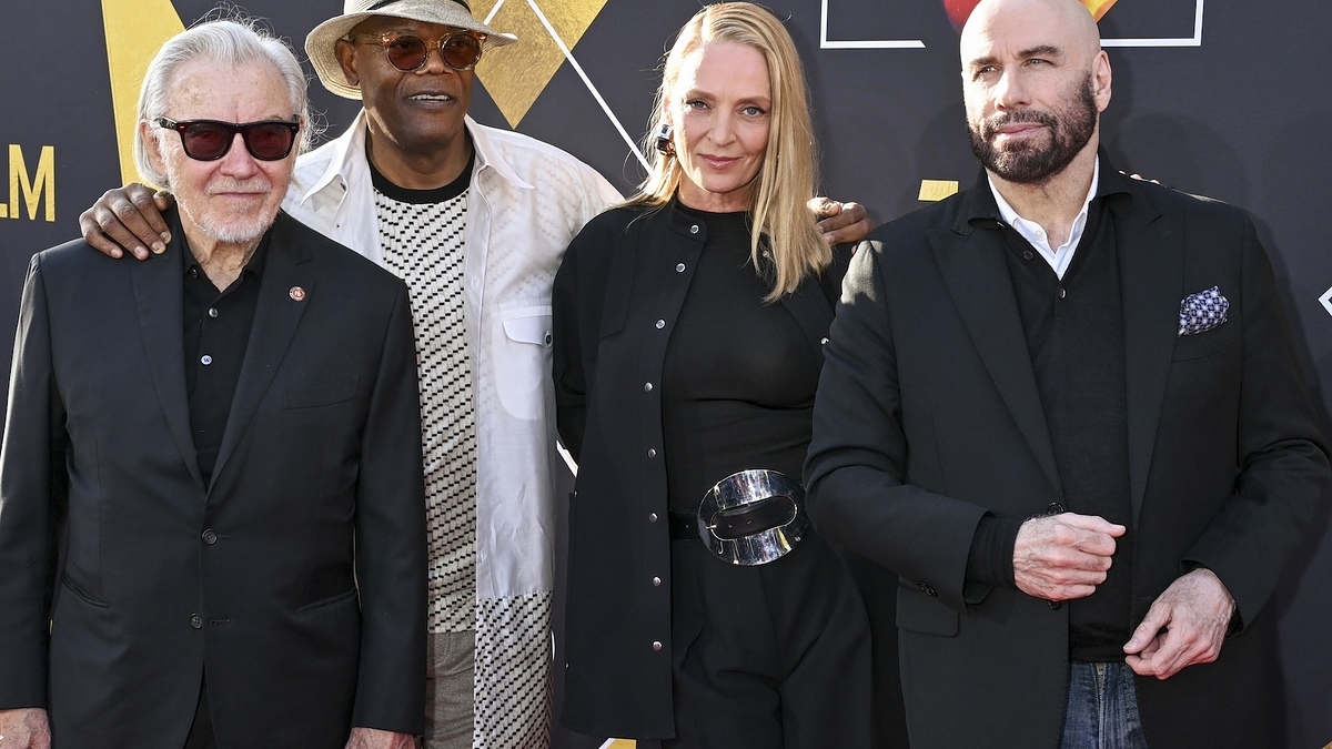 Four individuals posing together at an event; two men wear suits, one woman in a dress with a waist belt, and one man in a white outfit with a hat