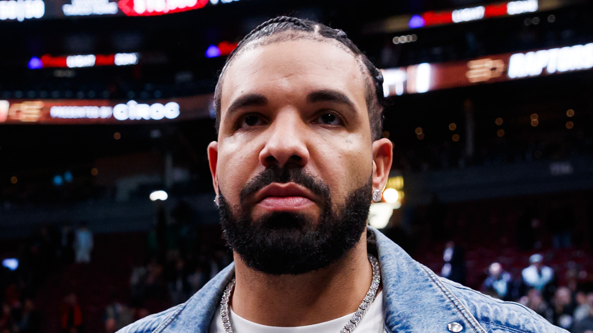 Drake wearing a denim jacket at a sports event, looking into the camera