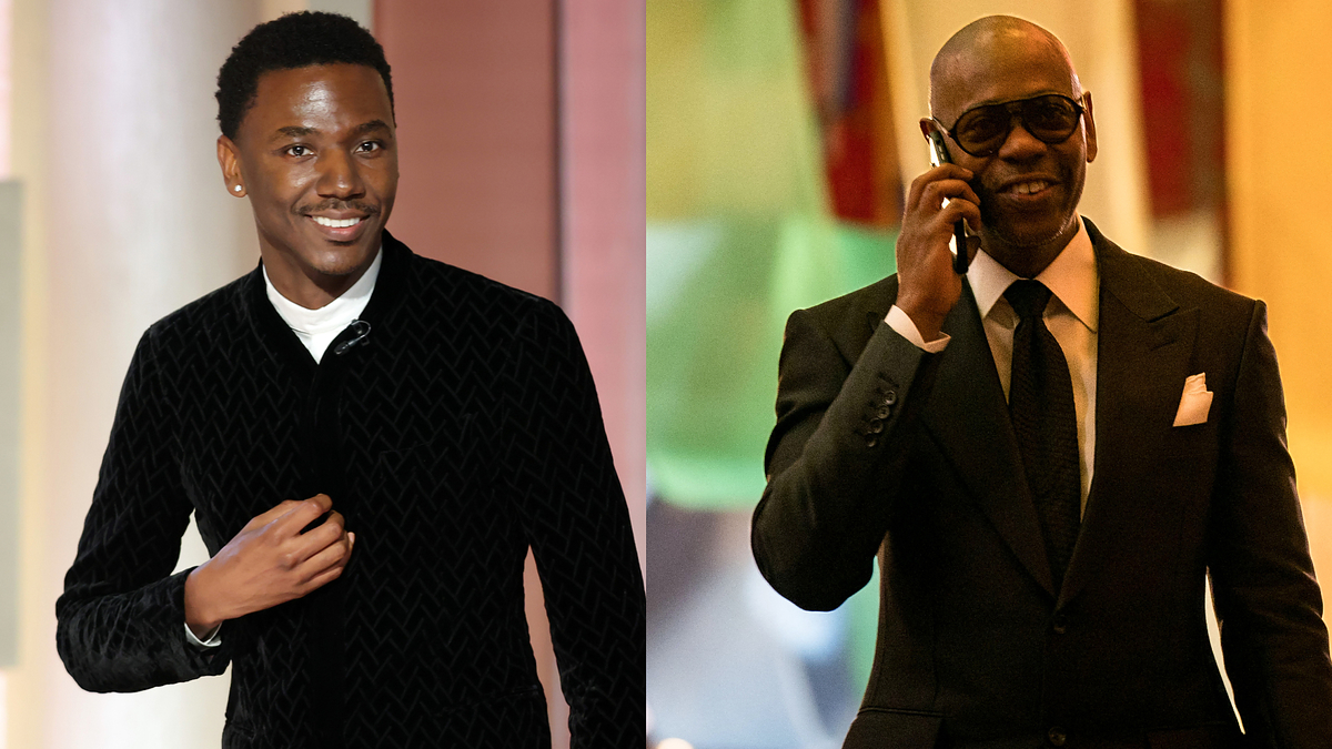 Two men at an event; one in a patterned jacket over a shirt, the other in a suit with sunglasses, using a phone