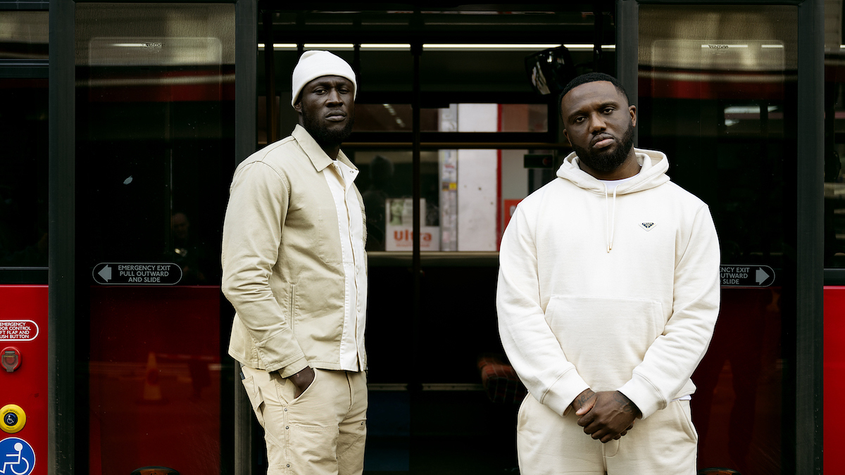Two men in casual attire stand in front of a bus, one in a beanie and the other in a hoodie
