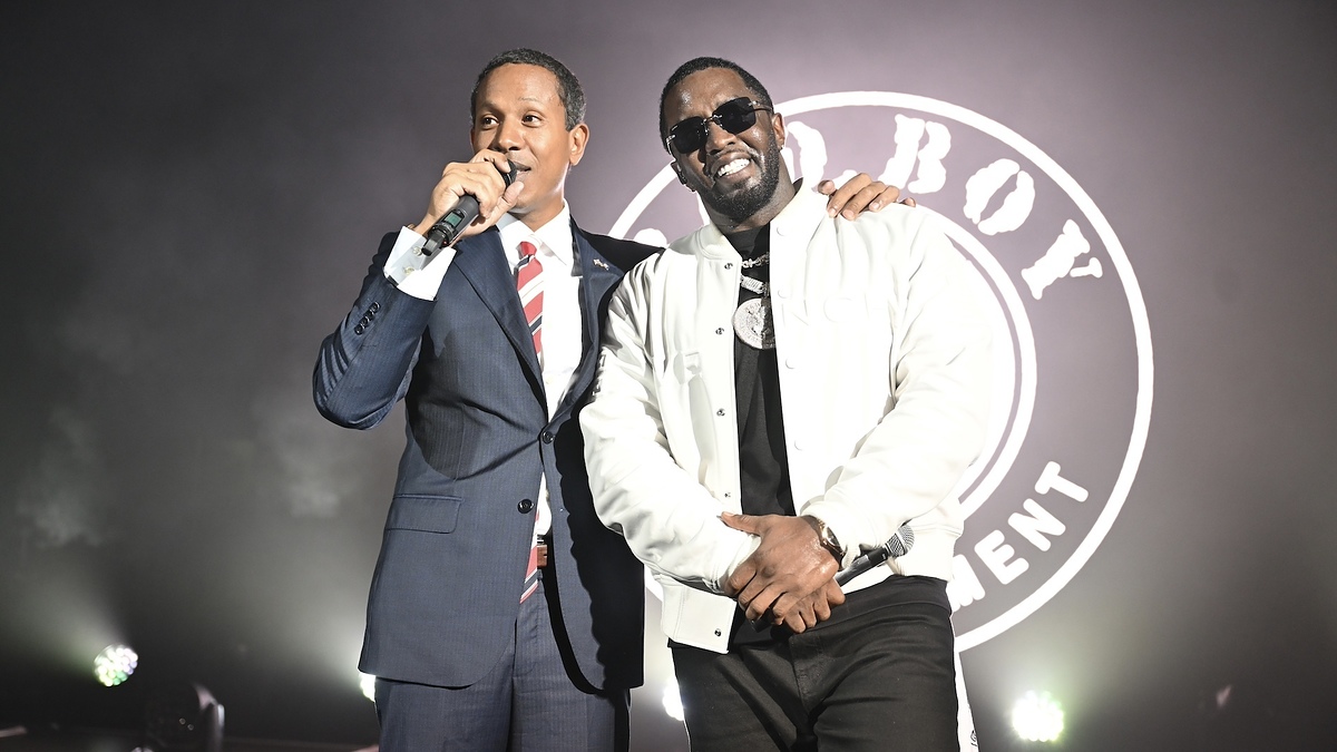 Two men on stage, one in a suit with a microphone, other in a jacket. They smile and embrace warmly