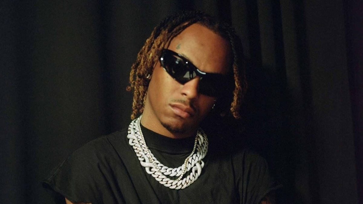 Music artist posing confidently with arms crossed, wearing sunglasses, a chain necklace, and a graphic tee