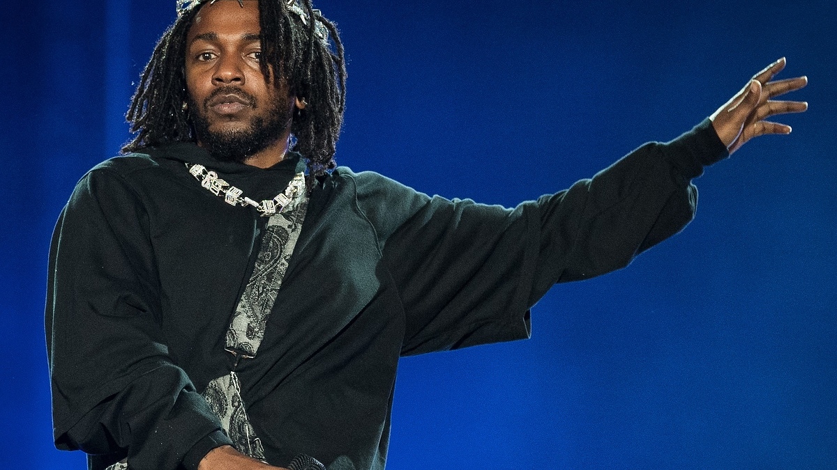 Hip-hop artist on stage in a casual black outfit with a microphone, performing with one arm extended