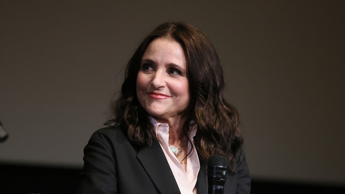 Woman seated with microphone, wearing a blazer and blouse, engaging in a discussion