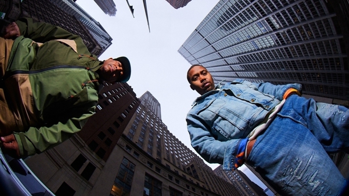 Two men, Nas and DJ Premier, sitting on a curb with skyscrapers rising around them, promoting "Define My Name"