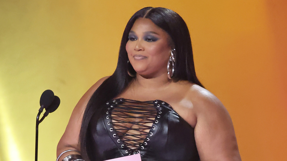 Lizzo stands at a podium with a microphone, wearing a laced-up outfit and earrings