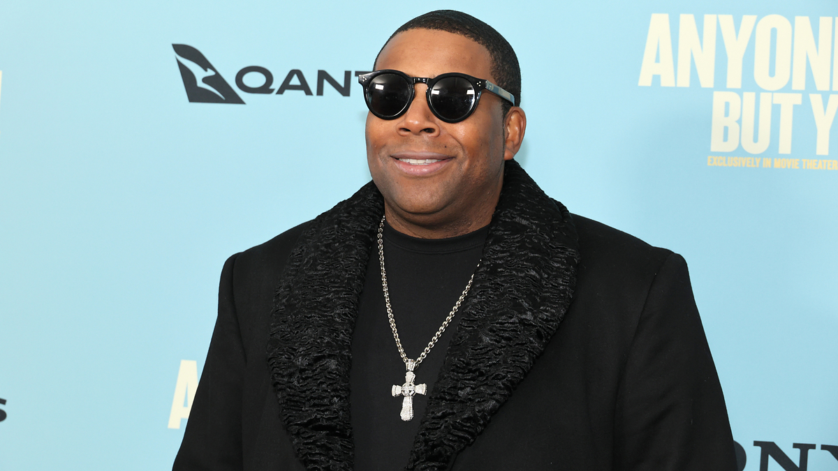 Man in a black jacket with fur trim smiling at a camera on the event backdrop