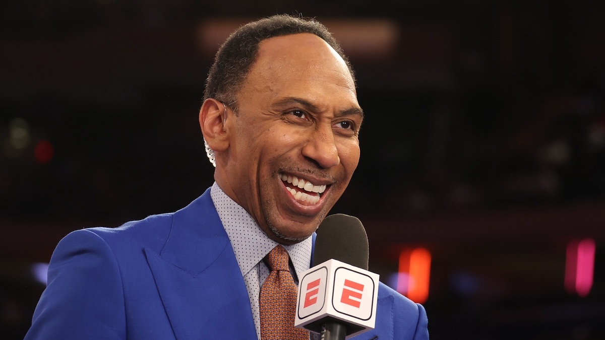 Stephen A. Smith wearing a blue suit, smiling while holding a microphone with ESPN logo