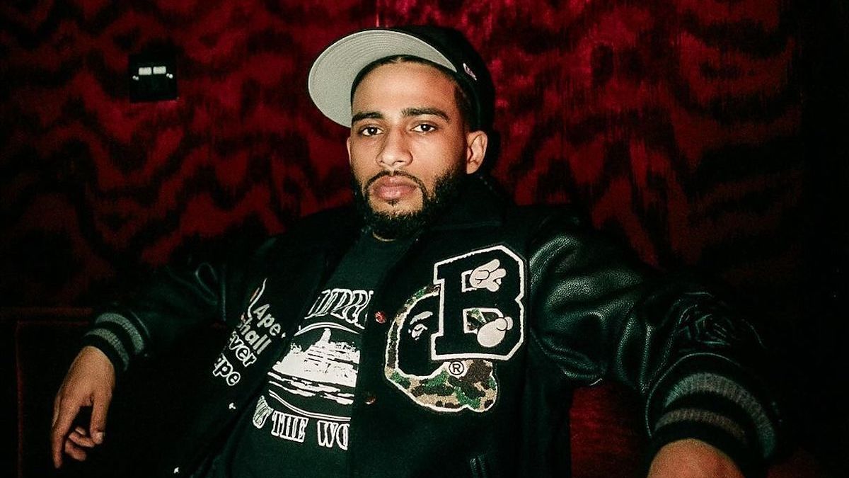 Man in a varsity jacket with embroidered patches and a baseball cap, posing in front of a textured backdrop