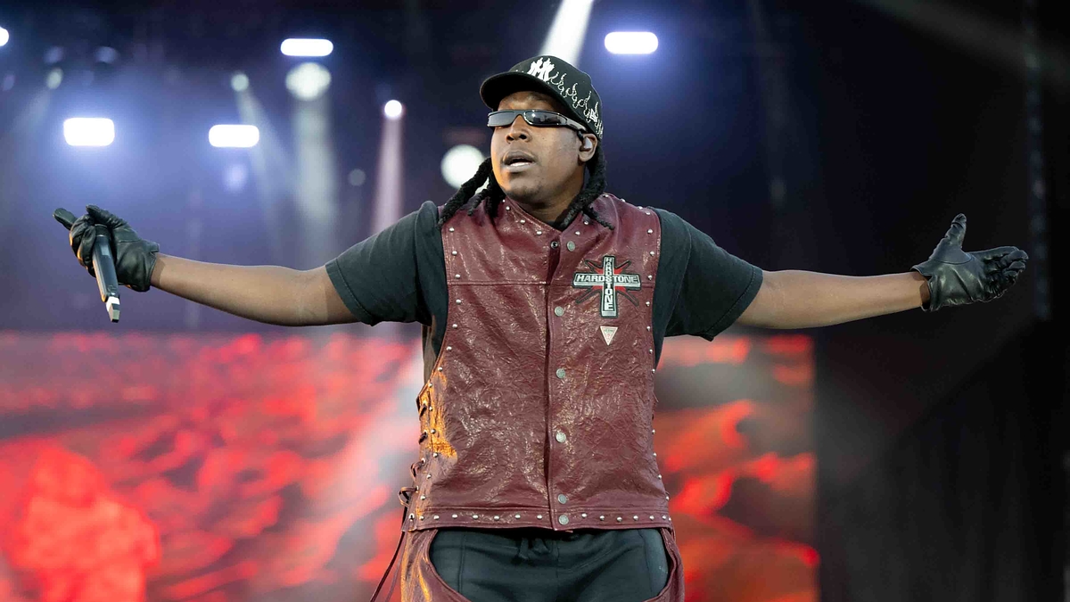 Music artist in a maroon vest and black cap performs on stage with arms outstretched