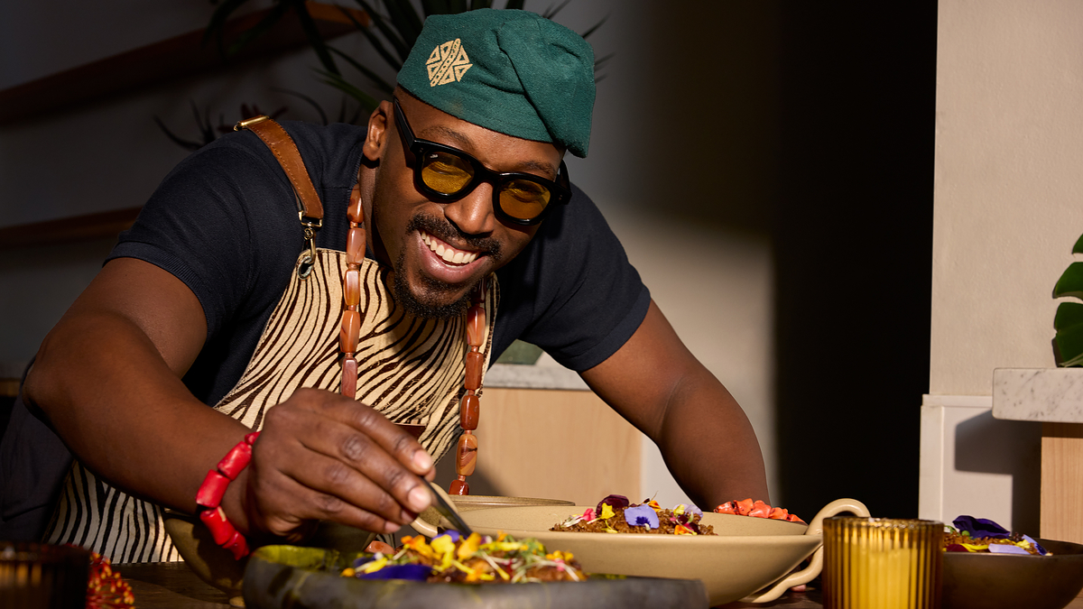 Man in apron and hat smiling while garnishing food on a plate at a table