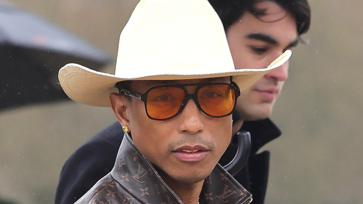Person in a wide-brimmed hat and sunglasses, sporting a patterned jacket. Another individual partially visible behind