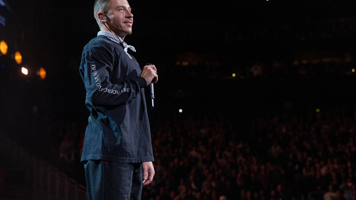 Man on stage speaking to an audience, wearing a jacket with text on the sleeve