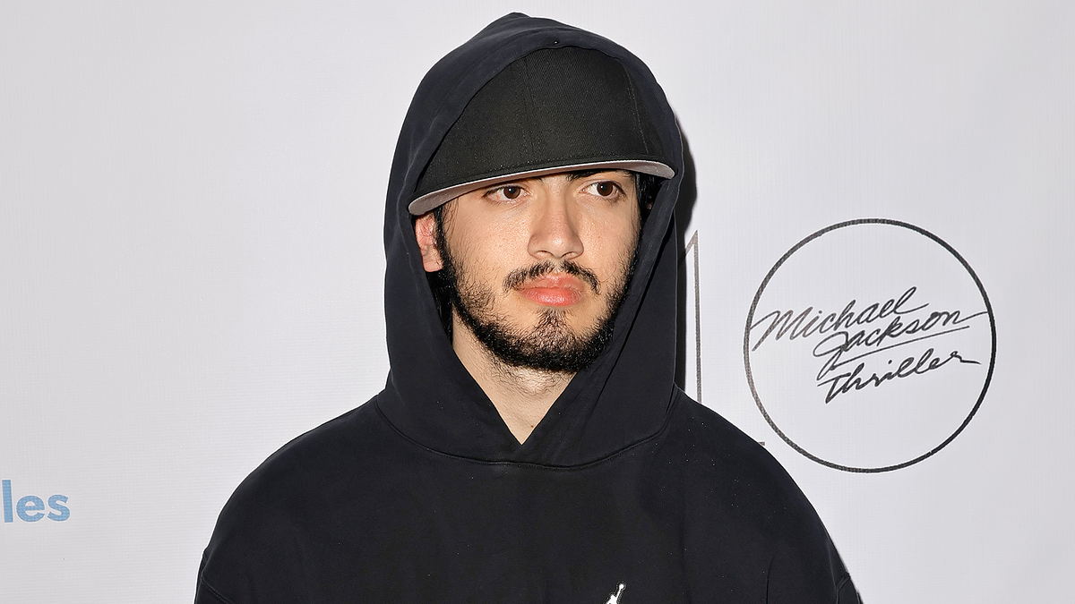 Man in a hooded black outfit standing in front of a promotional backdrop