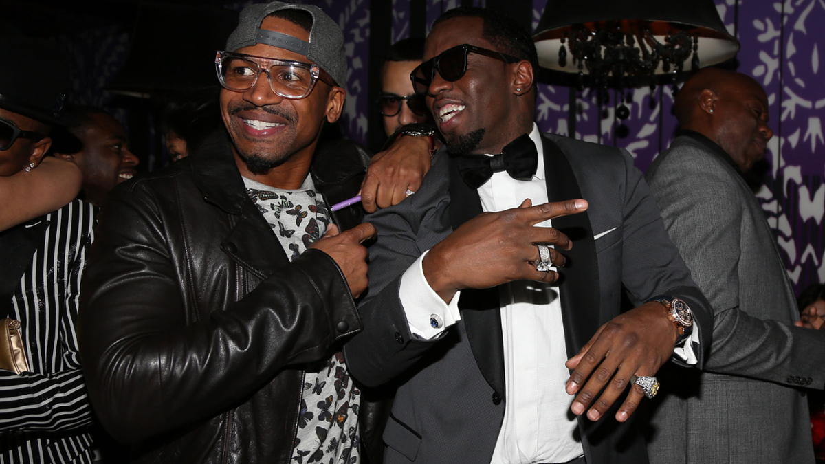 Two men laughing and embracing at an event, one in a leather jacket and the other in a tuxedo