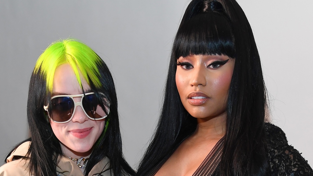Billie Eilish in oversized beige outfit, sunglasses, with Nicki Minaj in embellished black dress, both on event backdrop