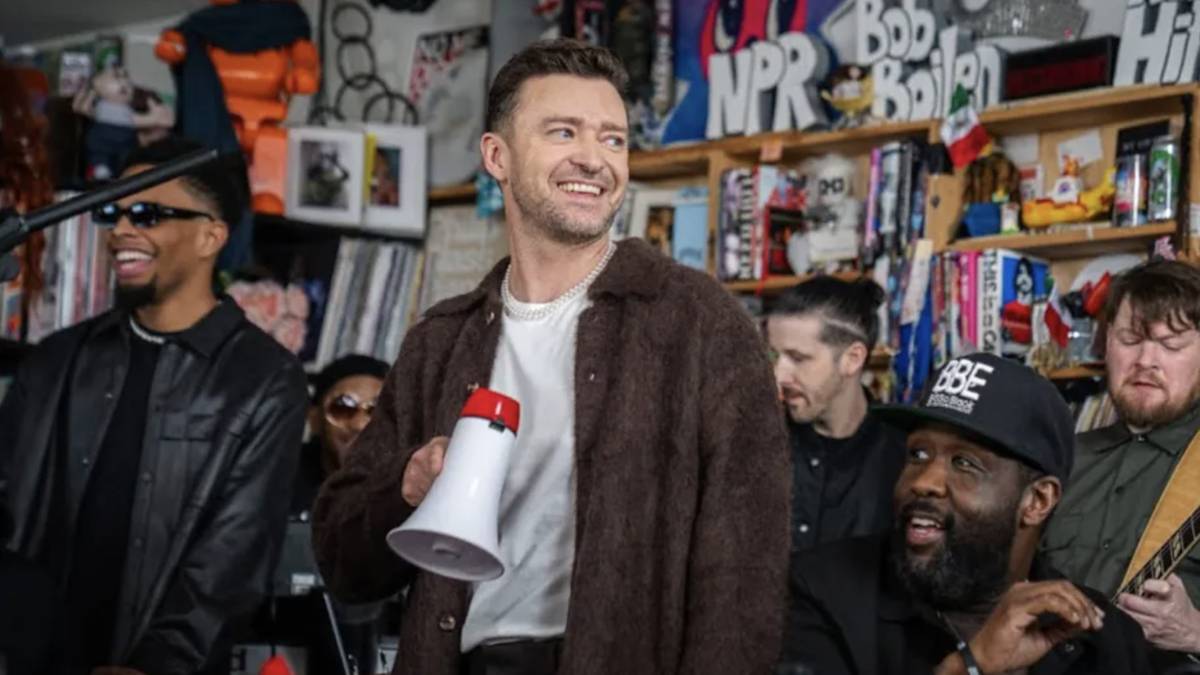 Justin Timberlake with a megaphone, flanked by a band in a room with eclectic decor