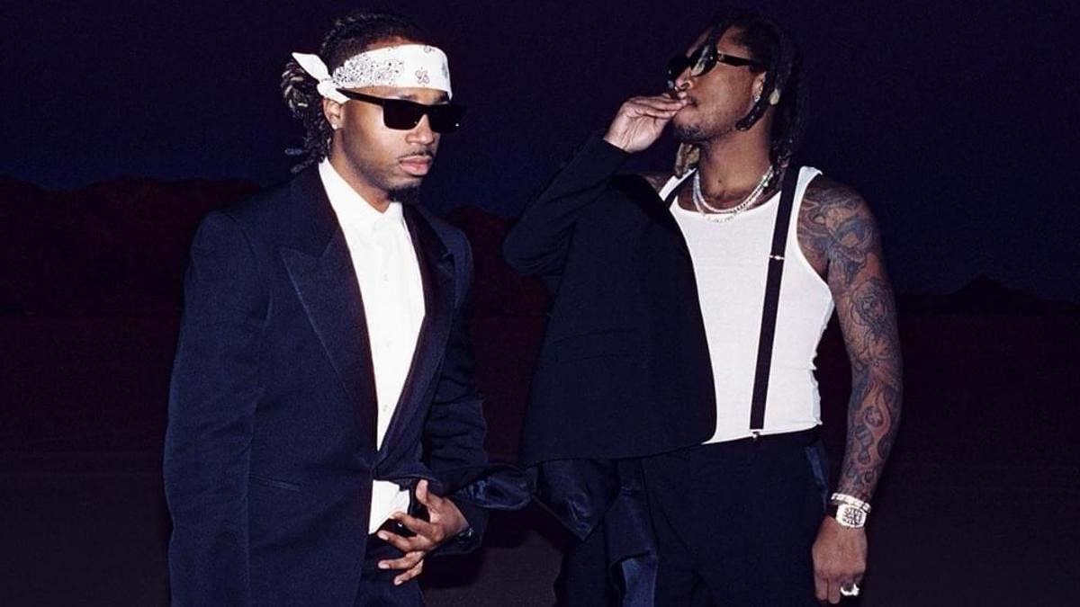 Two men in stylish suits stand confidently, one with sunglasses and a headscarf, the other with visible tattoos