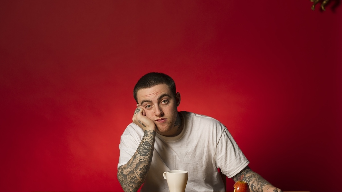 Person leaning on table with a mug & fruit, looking at camera, tattooed arms, plain background