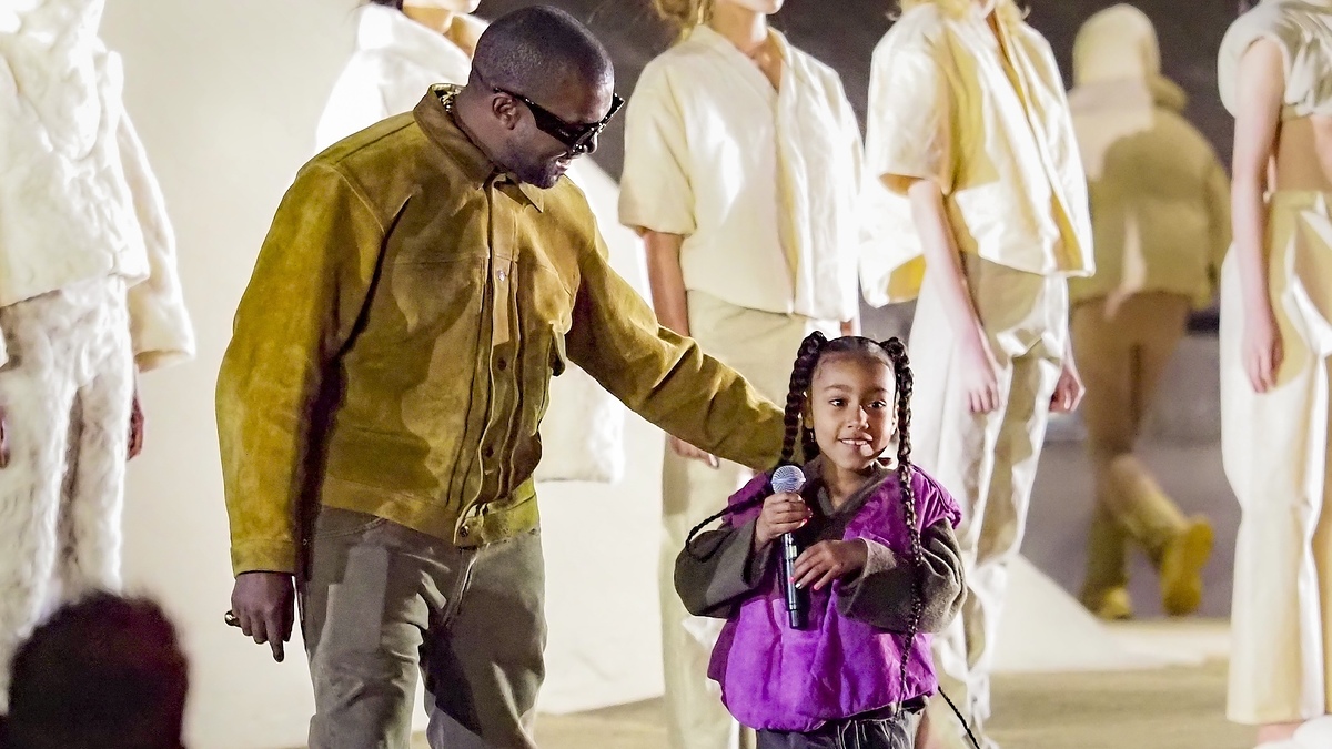 Man in a suede outfit holds the hand of a girl in a purple top and skirt, with models in background