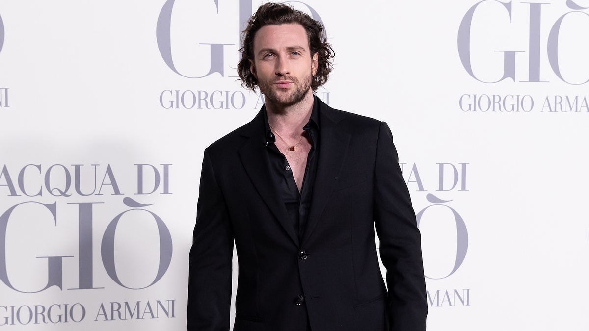 Man in a black suit without a tie standing in front of a promotional backdrop