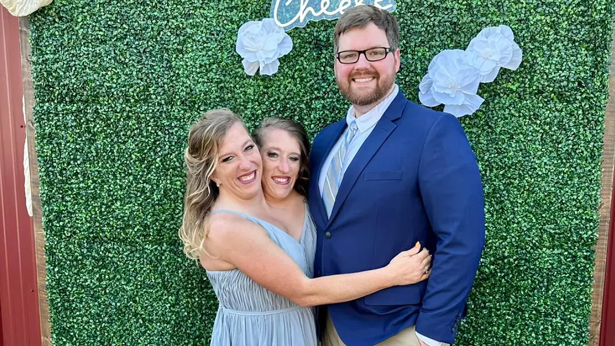 Three people smiling, one in a blue suit, flanked by two in dresses, in front of a greenery backdrop with white decorations