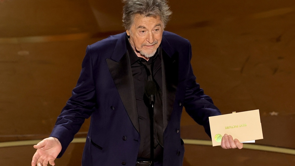Man in a navy suit holding an envelope on stage with a microphone, expressing an animated gesture