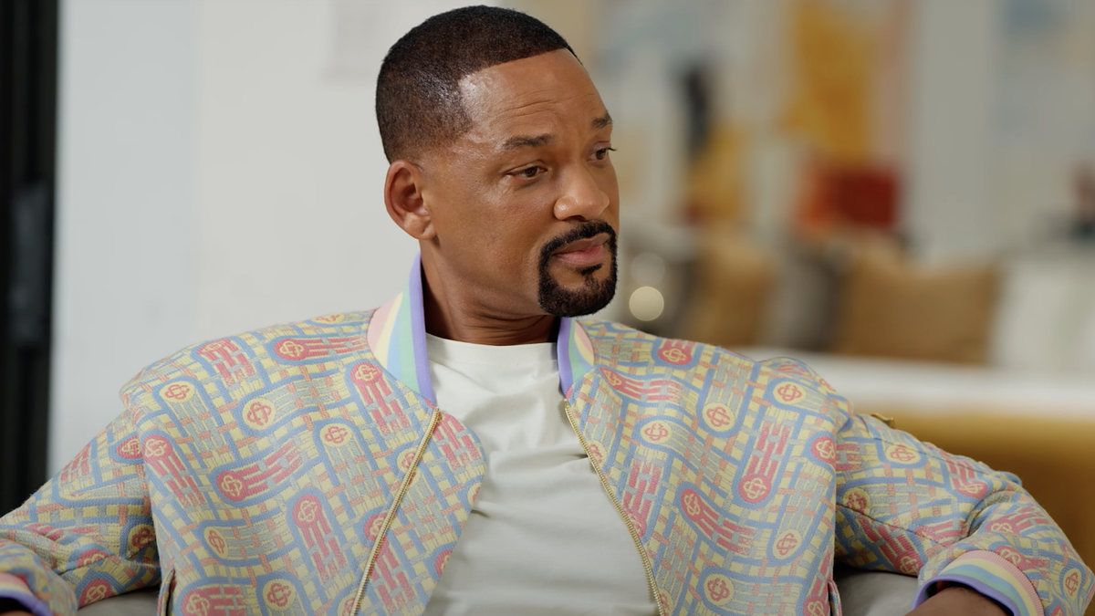A man in a printed suit sits on a couch in an interview setting, looking to the side