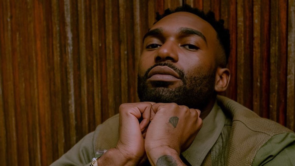 Man with beard posing with hands clasped, wearing layered shirts, heart tattoo on hand