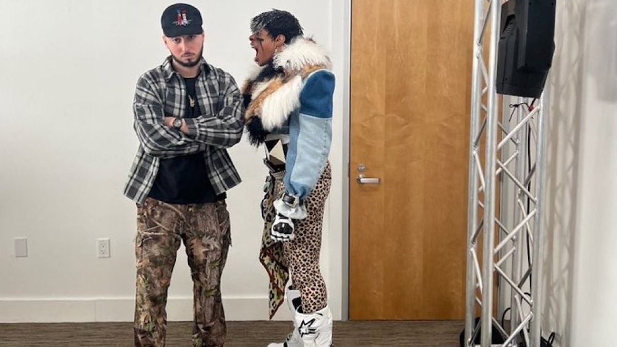 Two people in eclectic outfits standing in a room, one in a fur-collared jacket and animal print pants, the other in camo pants and a cap