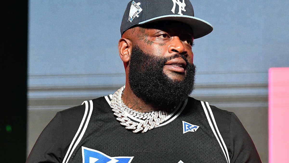 Man in sports cap, diamond necklace, and logo-printed shirt standing on stage.
