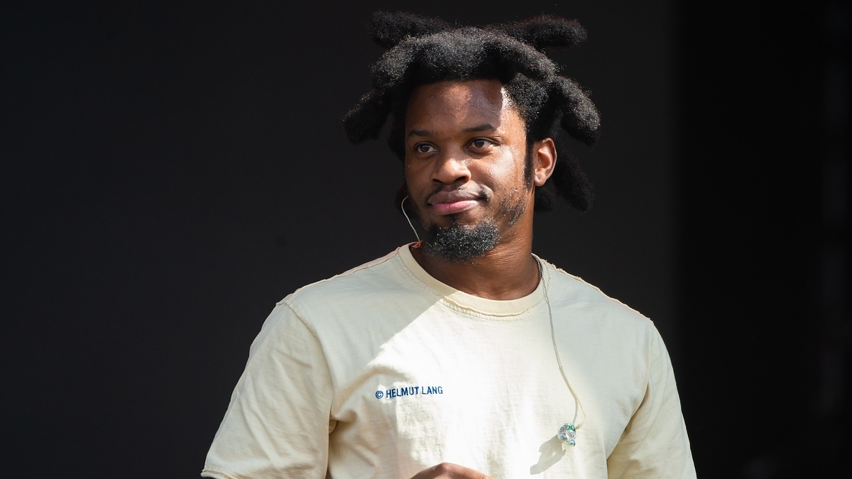 Man with unique hairstyle, wearing a Helmut Lang tee, and earphones, listening intently