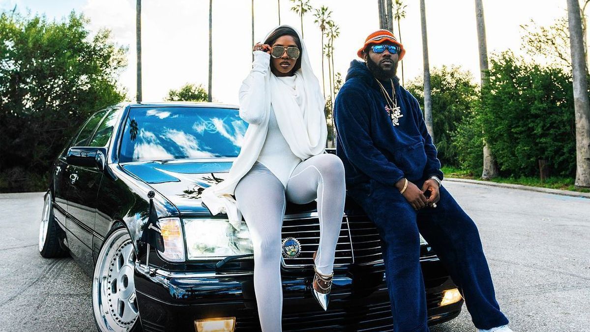 Two musicians pose by a car with "100 MILLION" text, one standing in white outfit, one sitting in blue, both with sunglasses