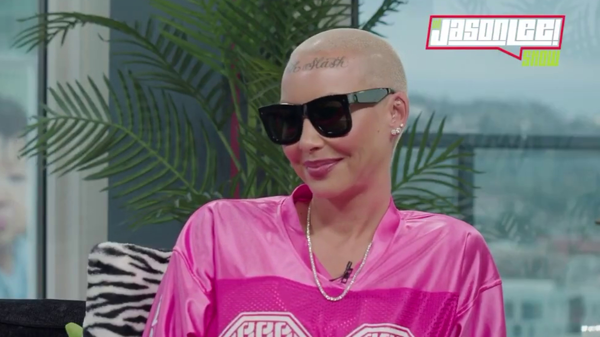 Amber Rose in sunglasses, buzz cut with tattoo, wearing a pink sports jersey, smiling during an interview