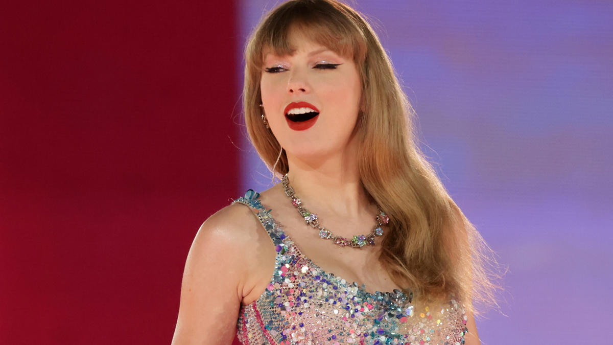 Taylor Swift is on stage singing, wearing a sparkly dress with a necklace