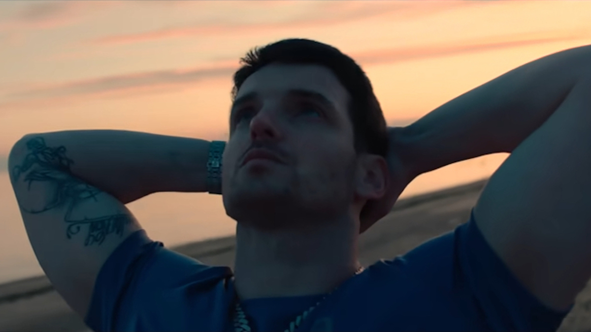 Man with tattoos on left arm gazes upwards, hands behind head, with sunset in background