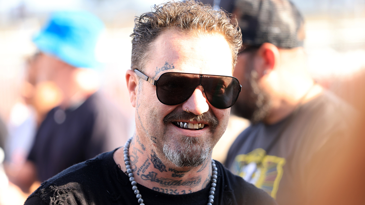 Man with sunglasses, tattoos, and facial hair smiling outdoors