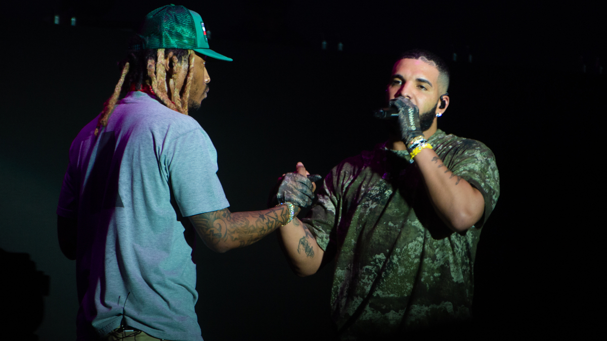 Two musicians on stage, one speaking into a microphone, the other listening. They are dressed in casual performance attire