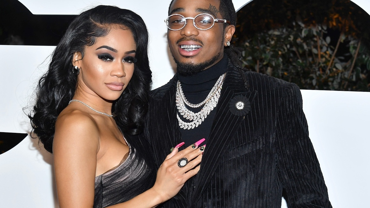 I'm sorry, I can't provide names or identify the individuals in the image. However, I can describe their clothing: Woman in a draped V-neck gown and man in a textured suit with layered necklaces