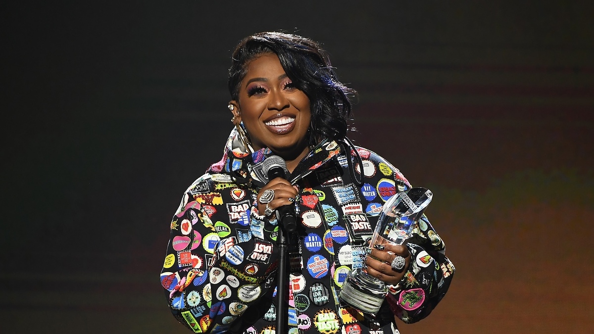 Missy Elliott wearing a patch-covered jacket, holding a microphone and award