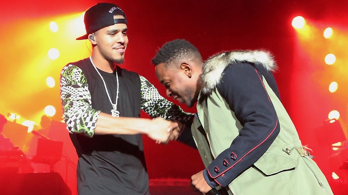 Two artists on stage, one in a printed jacket and cap, the other in a coat with red detailing, shaking hands